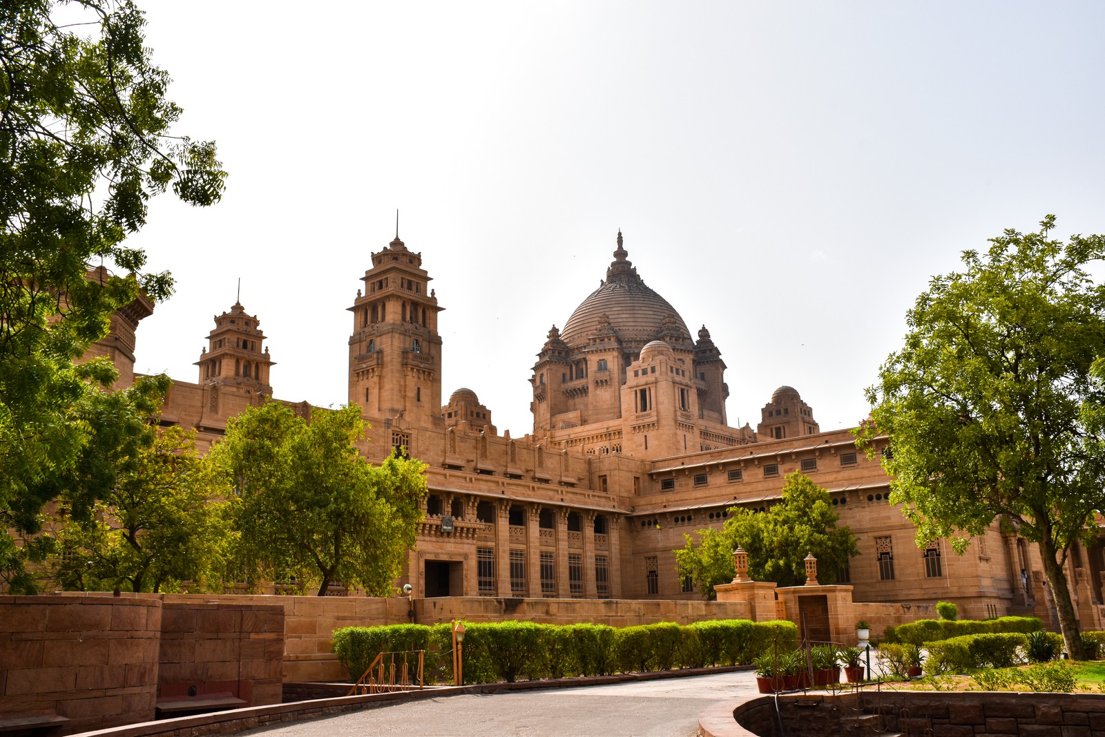 An angled exterior view of Umaid Bhawan Palace’s dome and sandstone towers, framed by courtyard trees.